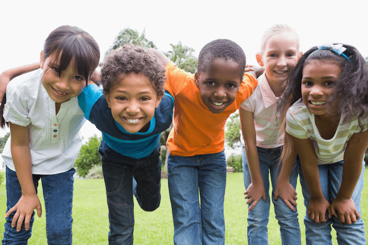 A group of five smiling children, diverse in appearance, lean forward together on a grassy field.